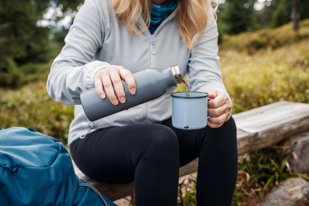 Woman pouring tea drink from flask into travel mug. Refreshment during hiking in the forestの写真素材
