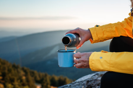 Woman tourist pouring hot drink from flask into travel mug in mountains. Refreshment during hiking outdoorsの写真素材