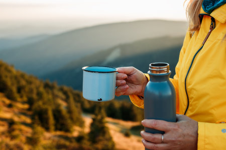 Refreshment after successfully climbed mountain summit. Female hiker drinking hot drink from travel mug and enjoying view at mountains during sunsetの写真素材