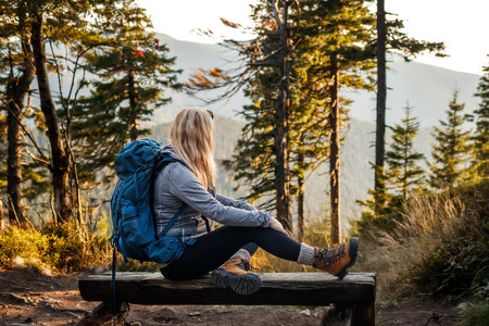 Relaxation during hiking in the mountain. Woman with backpack resting on wooden bench in forest. Suitable place for camping in the mountainsの写真素材