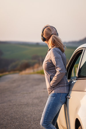 Woman with headphones leaning at car and resting during a road trip. Female person enjoying solo travelの写真素材