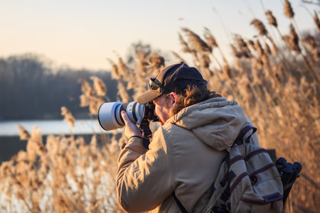 Photographer with camera hiding behind reeds at lake and photographing wildlife. Leisure activity in nature at sunriseの写真素材