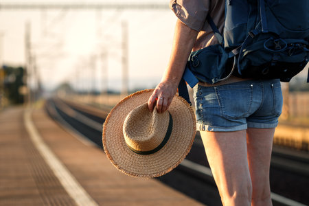Ready for summer journey. Traveler with backpack and hat waiting for train at railway stationの写真素材