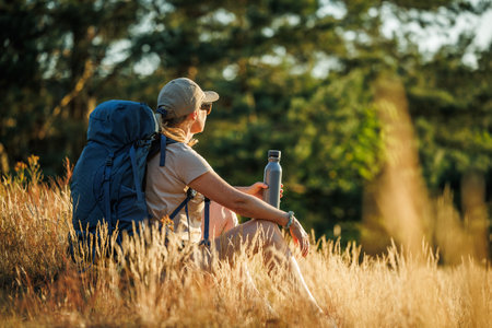 Woman with backpack is resting in forest and enjoying summer sunset. Relax while hiking on a long trekの写真素材