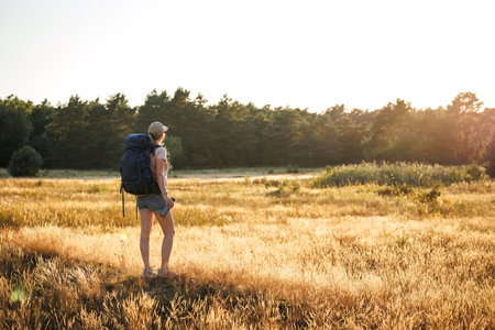 Female hiker with backpack and thermos is resting in nature and enjoying summer sunset. Woman relaxing while hiking on a long trekking trailの写真素材