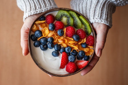 Woman holding healthy breakfast with fruits in coconut bowl. Yogurt with corn flakes and blueberry, strawberry, kiwi and raspberry. Vegetarian foodの写真素材