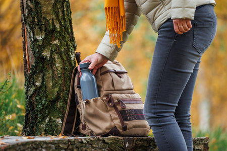 Hiker taking flask out of backpack to refresh with hot drink during hiking in autumn forest. Trekking in woodland at fall seasonの写真素材