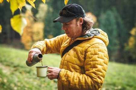 Hiker with yellow jacket is pouring hot drink from flask into travel mug in autumn forest. Resting and refreshment during hiking outdoorsの写真素材