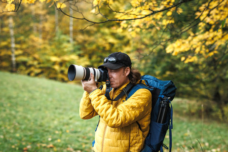 Hiker with backpack is wearing yellow jacket and photographing at his camera. Nature or landscape photographer in autumn forestの写真素材