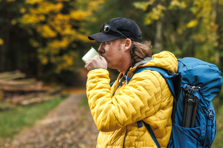Hiker with backpack and yellow jacket drinking hot tea from travel mug in autumn forest. Refreshment and resting during outdoor hikingの写真素材