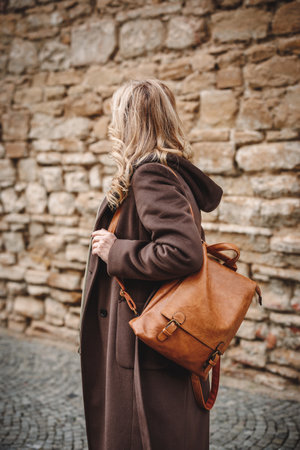 Stylish woman in brown coat with leather backpack walking next to stone wall at city street. Trendy warm clothing and fashion accessoriesの写真素材