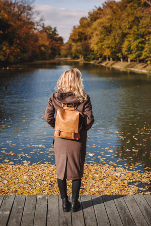 Stylish woman in brown coat with leather backpack standing at lake in autumn park. Trendy warm...