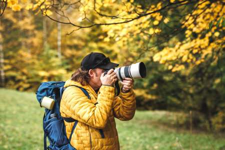 Hiker with backpack is wearing yellow jacket and photographing at his camera. Wildlife photographer in autumn forestの写真素材