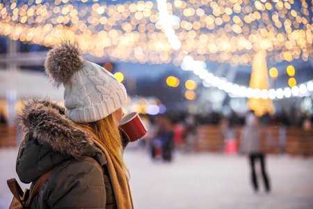 Woman looks at people skating on ice rink and drinks mulled wine or punch. Christmas market and...