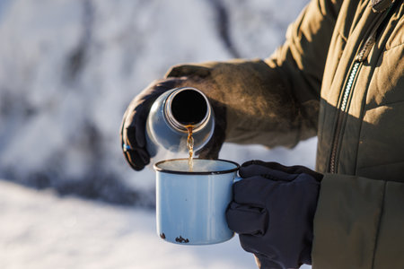 Hiker pouring hot drink from flask into travel mug. Refreshment during winter trekking in cold weatherの写真素材