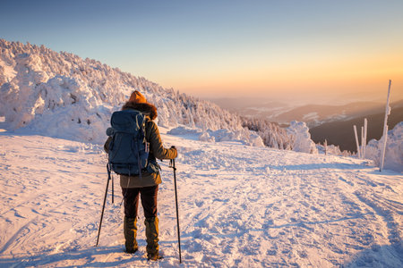 Woman with backpack and Nordic walking poles trekking in cold weather. Hiking in snow at winter mountain during sunset. Sports and outdoor seasonal activitiesの写真素材