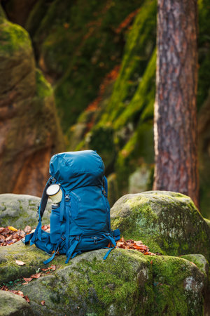 Backpack with travel mug on rock in forest. Hiking and trekking equipment. Essentials for outdoor sportsの写真素材