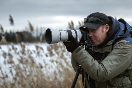 Wildlife photographer is setting up camera on tripod outdoors. Man photographing landscape or animals at lake in winter. Bird watchingの写真素材