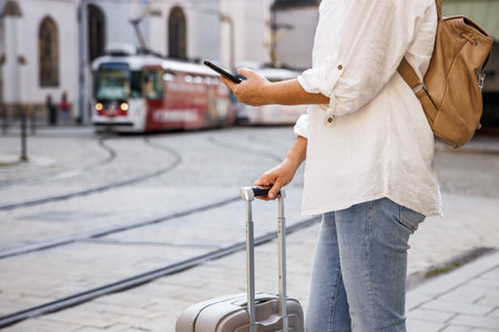 Woman tourist with suitcase and smart phone waiting for tram on street. Travel by public transportation in the cityの写真素材