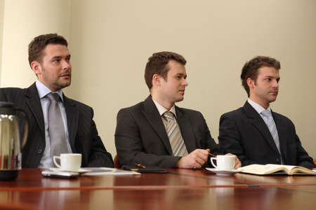 Three businessmen in suits sitting at a conference table, taking part in a meeting and/or...