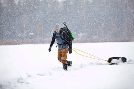 winter condition training - man with backpack walking pulling tyre on snowy field in snowstormの写真素材