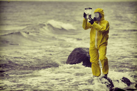 Specialist in protective suit checking sample of water in container on rocky sea, ocean shoreの写真素材