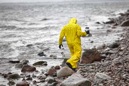 Scientist in protective suit with plastic container walking in on rocky beach - back view