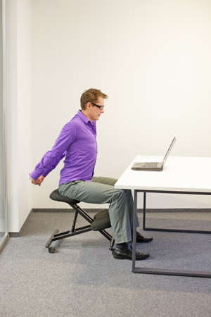 male office worker sitting on kneeling stool, stretching arms during short break in work at his desk in the officeの写真素材