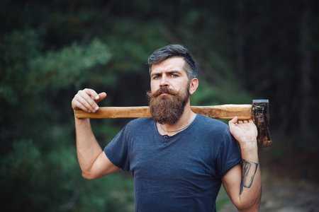 Brutal brunette bearded man in warm hat with a hatchet in the woods on a background of treesの写真素材