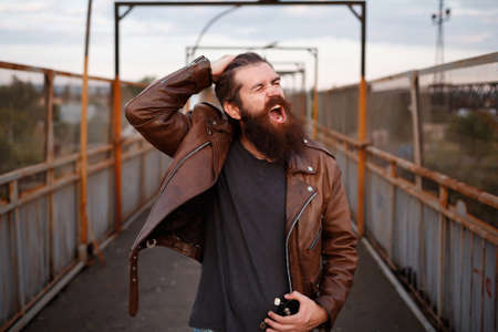 A stern brutal man with a long mustache in a brown leather jacket holds his hair and shouts against the background of a pedestrian crossing over the railwayの写真素材