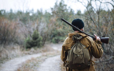 A bearded man-hunter in a black hat, in a hood, in a khaki jacket with a gun on his shoulder and cartridges on his chest smokes a pipe against the background of the forestの写真素材
