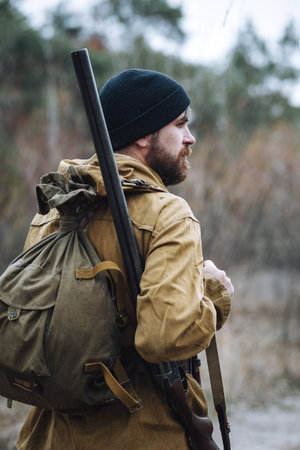 a bearded brutal hunter man in a dark warm hat, khaki jacket with a gun on his shoulder shaves his...