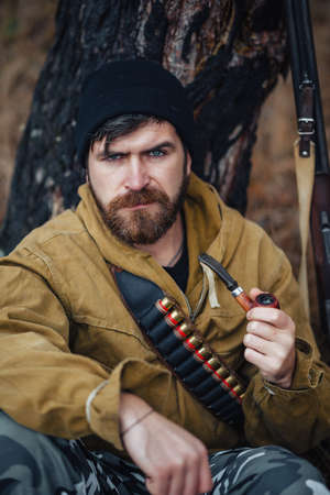 a bearded brutal hunter man in a dark warm hat, khaki jacket with a gun on his shoulder shaves his...
