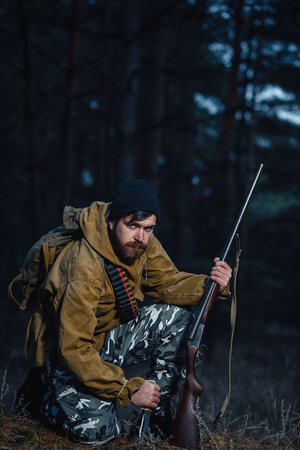 An experienced male hunter with a beard in a green khaki jacket and a long cloak holds a gun in his...