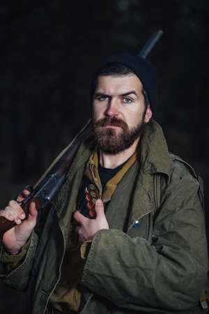 Serious brutal bearded man hunter in a dark hat and khaki jacket in a long cloak holds an unloaded...