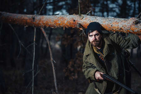 Old bearded man hunter in a warm hat, in a khaki jacket with a hunting rifle and a large backpack with cartridges against the background of a dark forest at nightの写真素材
