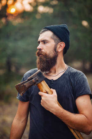 a bearded man in a hat and a dark sleeveless T-shirt with a large mustache and gray hair with an ax on his shoulder walks along a country road in the forestの写真素材