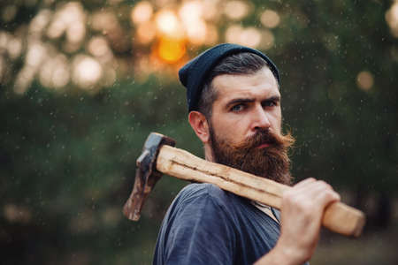 a bearded man in a hat and a dark sleeveless T-shirt with a large mustache and gray hair with an ax on his shoulder walks along a country road in the forestの写真素材