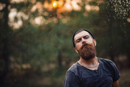Old man with a large mustache in a dark sleeveless T-shirt and in a warm hat with gray hair shaves his beard with an old rusty metal ax against the backdrop of a sunset forestの写真素材