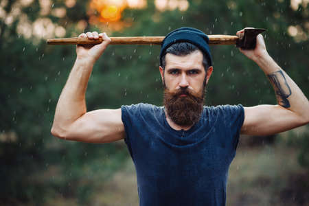 Old man with a large mustache in a dark sleeveless T-shirt and in a warm hat with gray hair shaves his beard with an old rusty metal ax against the backdrop of a sunset forestの写真素材