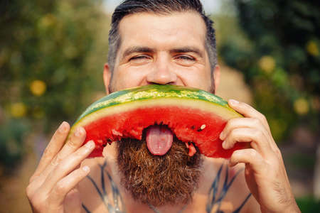 Funny tanned bearded man grimaces and holds on a long tongue a piece of ripe bitten watermelon in the rain in a green gardenの写真素材