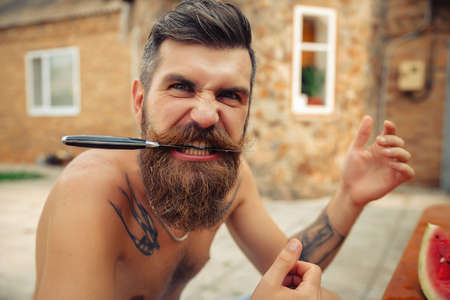 Wet tanned bearded man with a big red mustache laughs and holds a piece of ripe red watermelon in the rain in a green gardenの写真素材