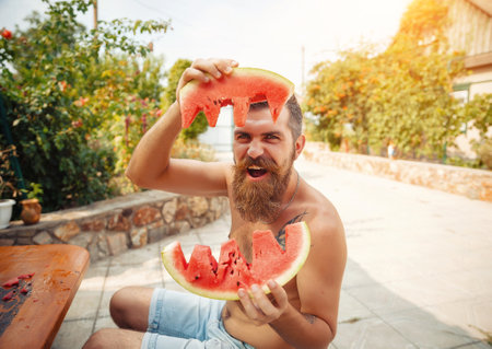 Wet tanned bearded man with a big red mustache laughs and holds a piece of ripe red watermelon in the rain in a green gardenの写真素材