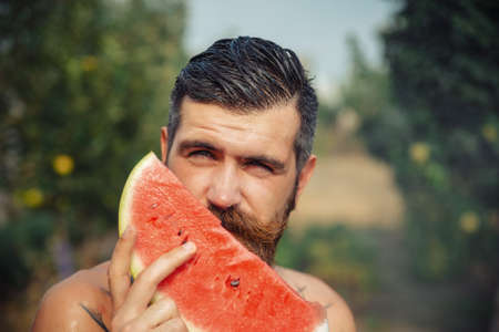 A man with a stylish hairstyle, a long beard and a tattoo with great pleasure eats watermelonの写真素材