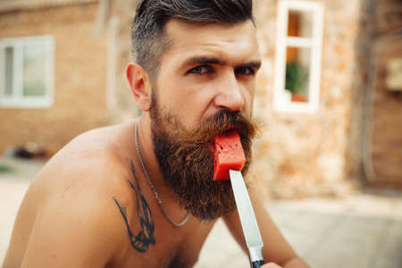 Wet tanned bearded man with a big red mustache laughs and holds a piece of ripe red watermelon in the rain in a green gardenの写真素材