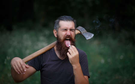 a man lumberjack with a long curly beard in a warm hat and a dark T-shirt with an iron ax on his shoulders against a background of a dark forestの写真素材
