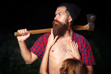 A brutal bearded man lumberjack in a hat and a checkered red shirt with an ax holds a blonde girl in a bra behind her assの写真素材