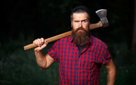 Bearded man lumberjack in warm hat and checkered red shirt with iron ax on his shoulder against dark...