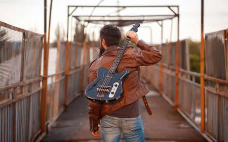 A bearded rock band guitarist with gray hair in a brown leather jacket and blue jeans with a black electric guitar lies on the ground by a fence at sunset and straightens his hairの写真素材