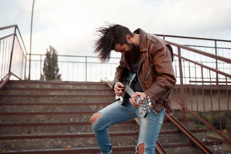 Mad rock guitarist with gray hair and a big beard with a mustache in a brown leather jacket and blue jeans plays a black electric guitar and screams against the backgroundの写真素材
